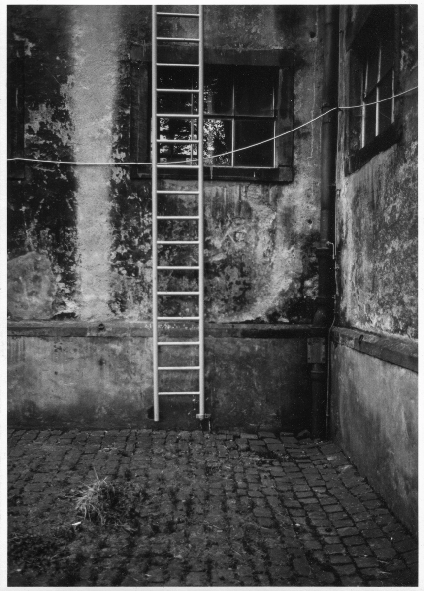 Schwarz-Weiß-Fotografie von Thomas Gaedtke:
Eine metallene Fluchtleiter hängt an einer verwitterten Hauswand in einem historischen Innenhof mit Kopfsteinpflaster. Ein offenes Fenster, bröckelnder Putz und urbane Verfall-Ästhetik prägen das Bild.